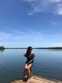 Full length of young woman looking at lake while sitting on pier against sky during sunny day