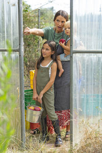Girl holding fruit bucket standing with mother carrying brother at doorway of greenhouse