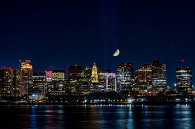 Illuminated buildings by river against sky at night