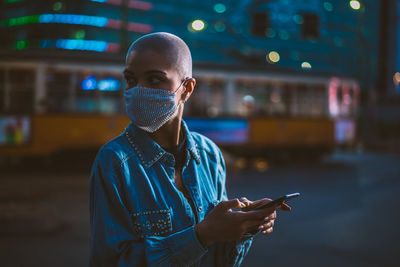 Young woman wearing mask with shaved head looking away while holding smart phone outdoors