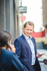 Smiling mature businessman with smart phone looking at coworker outdoors