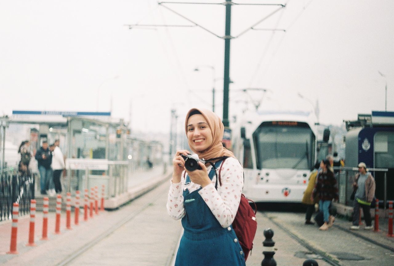 PORTRAIT OF YOUNG WOMAN STANDING BY TRAIN IN CITY