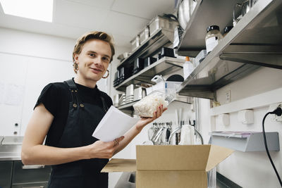 Portrait of smiling transgender owner with list and package in coffee shop kitchen