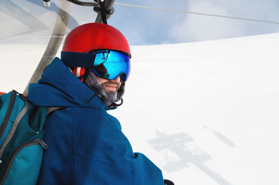 A skier in a helmet and a mask sits on a lift and smiles, against the backdrop of high snow-capped