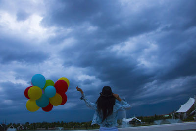 Rear view of woman holding helium balloons against cloudy sky