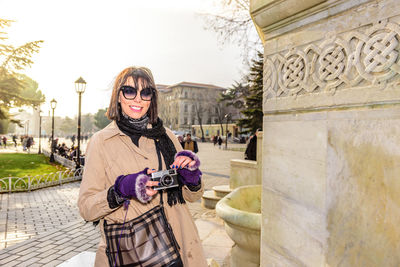 Portrait of woman holding camera while standing outdoors