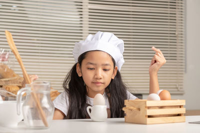 A little asian girl wearing a white chef hat is staring at a duck egg