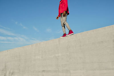 Young man in red jacket and checked pants walking on concrete wall