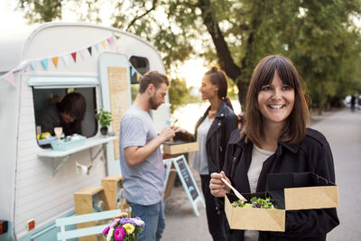 Smiling customer holding disposable salad box on street with friends and owner outside food truck in background