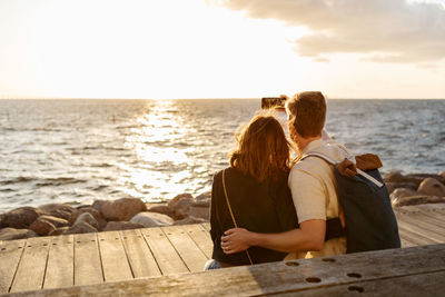 Rear view of couple taking selfie on smart phone while sitting against sea