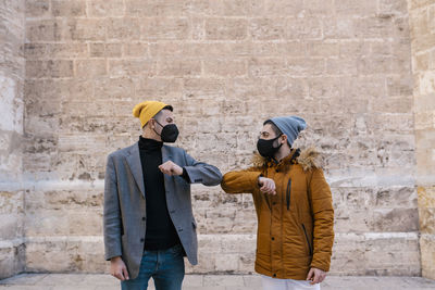 Young men wearing knit hat and protective face mask giving elbow bump while greeting against wall