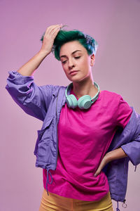 Front view of pensive millennial female with dyed hair and headphones wearing casual clothes with retro colors standing on purple in studio