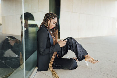 Young businesswoman with smart phone leaning on glass wall