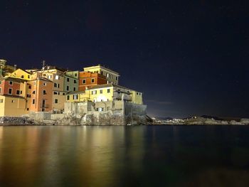 Illuminated buildings by sea against sky at night
