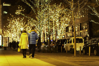 Rear view of couple walking on illuminated sidewalk at night