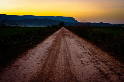 Dirt road amidst field against sky during sunset