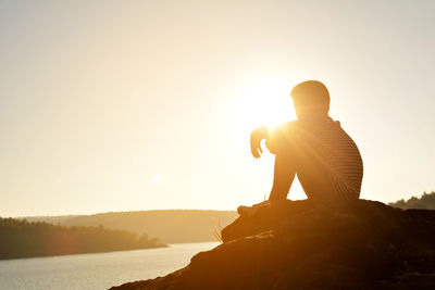 Silhouette teenage boy sitting on rock formation by lake against sky during sunset