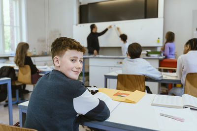 Portrait of smiling boy looking over shoulder while sitting on desk in classroom