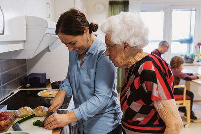 Side view of senior woman looking at daughter cutting zucchini in kitchen