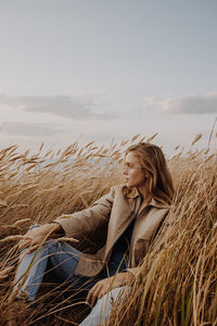 Young woman looking away while sitting by plants against sky