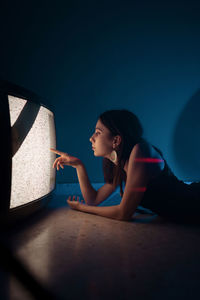 Side view of female model in black dress lying down on floor near glowing old television in dark studio