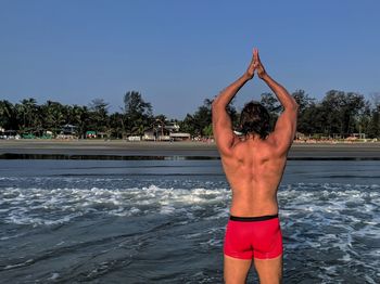 Rear view of shirtless man with hands clasped standing in sea against sky