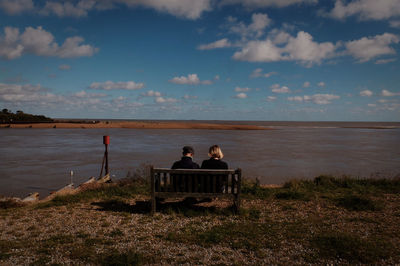 Rear view of man sitting on beach against cloudy sky