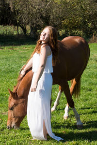 Side view of smiling young woman with horse standing on grassy field against trees