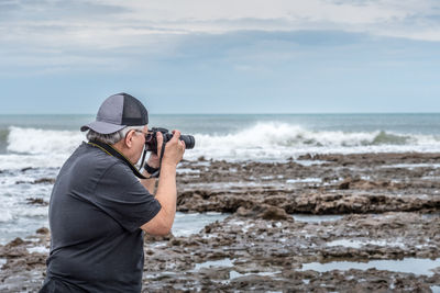 Half body rear view of mature adult male with cap taking a photo on the rocks near the sea.