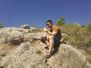 Full length of young man drinking water while sitting on rock during sunny day