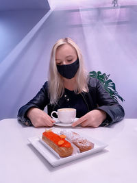 Woman sits in a cafe with a medical mask on her face and looks sadly at a cup of coffee and eclairs.