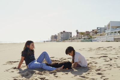 Side view of two girls sitting on beach and playing with dog