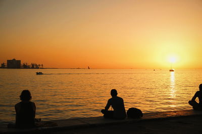 Silhouette men sitting on shore against orange sky