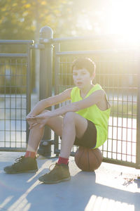 Portrait of boy sitting on basketball against fence