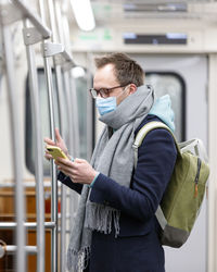 Side view of man wearing mask using smart phone while standing in subway train