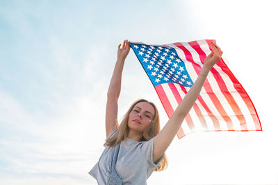 Young millennial blonde woman standing with american flag next to blue sky. flag of the united 