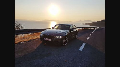 Car on road by sea against sky during sunset