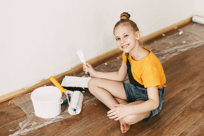 A girl in a denim overalls and a yellow t-shirt helps to paint the walls in an apartment white.