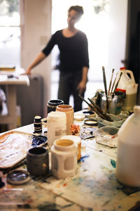 Paint bottles and containers on messy table with woman in background