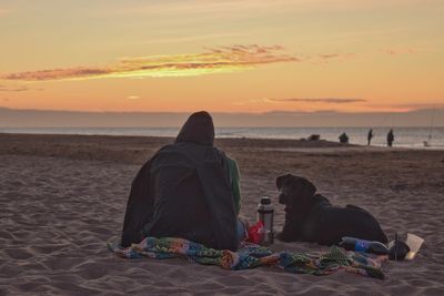 Rear view of men sitting on beach against sky during sunset