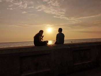 Couple sitting by sea against sky during sunset