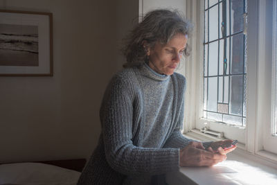 Young woman using mobile phone while sitting on window