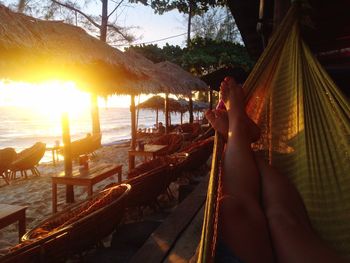 Low section of woman relaxing on hammock by parasols at beach during sunset