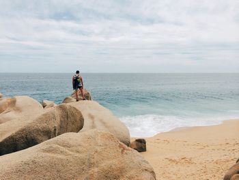 Rear view of mid adult man with backpack standing at beach against cloudy sky