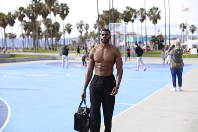 Portrait of shirtless muscular man holding bag while standing at basketball court