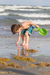 Full length of shirtless boy holding toy while bending at beach