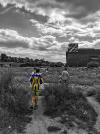 Rear view of people walking on dirt road against cloudy sky