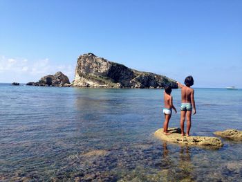 Shirtless siblings standing on rock formation while pointing at sea against clear blue sky