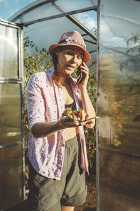 Farmer holding tomatoes talking on mobile phone at doorway of greenhouse