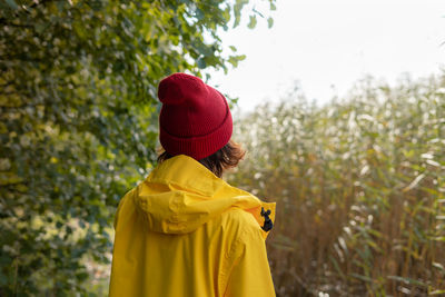 Back view of woman in bright yellow raincoat and red hat looks at reeds